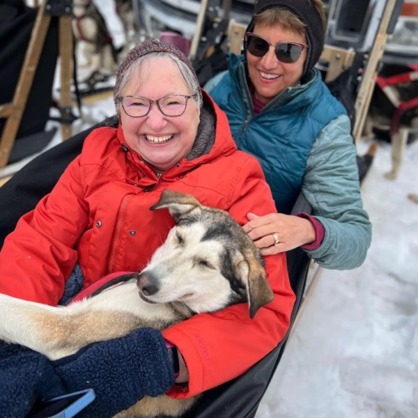 Two people in a sled with a dog, smiling in a snowy setting.