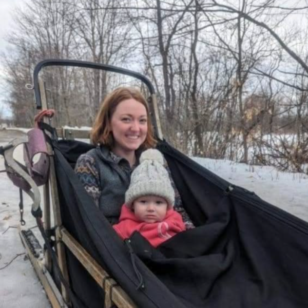 Woman and baby in a sled on snowy ground, surrounded by bare trees.
