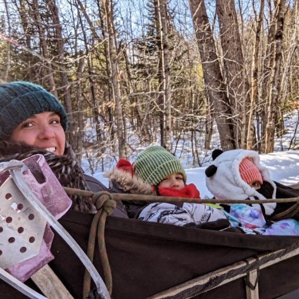 Three people bundled in winter clothing riding a sled through snowy woods.