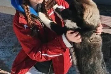 Person in red jacket and white hat kneeling and playing with a black and white dog in snowy area.