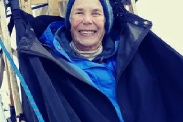 Person smiling while sitting in a sled on snow, wearing winter clothing and a beanie.