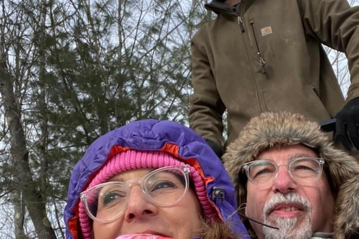 Three people bundled in winter clothing, one standing behind two sitting in snowy outdoor setting.