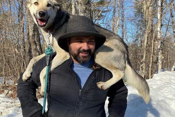 Smiling man in a hat with a dog on his shoulders in a snowy forest.
