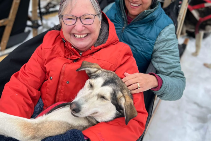 Two smiling people seated in a sled, holding a relaxed dog in snowy surroundings.