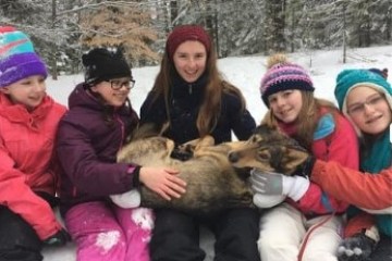 Five girls in winter clothing sitting in snow, holding a dog in a snowy forest.