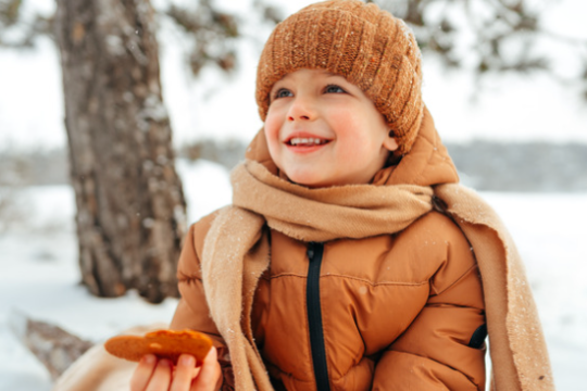Child in winter clothes sitting outdoors, holding a cookie, with snow-covered trees in the background.