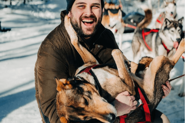 Man in winter clothes holding a sled dog in snowy landscape.